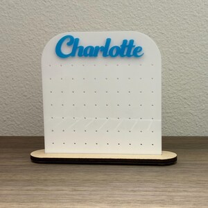 May include: A white acrylic earring display stand with the name "Charlotte" in blue script. The stand has multiple holes for earrings and sits on a wooden base. The overall design is simple and modern.