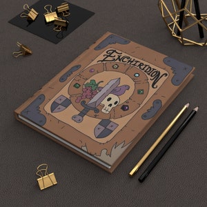 May include: A brown leather-bound journal with a whimsical illustration of a sword, skull, and grapes. The journal cover features the word "Enchiridion" in a stylized font.
