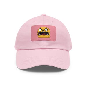 May include: A light pink baseball cap featuring a rectangular patch with a cartoon character's face. The character has yellow skin, black eyes, and an open mouth. The cap has a curved brim and a six-panel design.