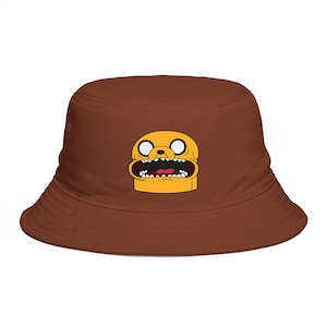 May include: A brown bucket hat featuring a cartoon character's face in yellow, black, and red. The hat has a wide brim and a rounded crown. The cartoon character has large white eyes, a wide open mouth, and a yellow body.
