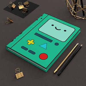 May include: A teal notebook with a cartoon face design, light blue screen, yellow cross, blue triangle, and red circle. Gold and black pencils and gold binder clips are also visible.