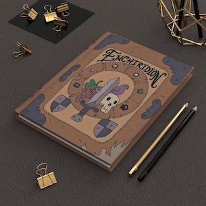 May include: A brown leather-bound journal with a whimsical illustration of a sword, skull, and grapes. The journal cover features the word "Enchiridion" in a stylized font.