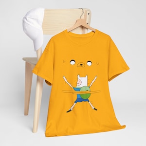 May include: Mustard yellow t-shirt with a cartoon illustration of Finn from Adventure Time. The design shows Finn with his arms outstretched, a green shield, and a portion of Jake's face above. A white baseball cap rests on a wooden chair.