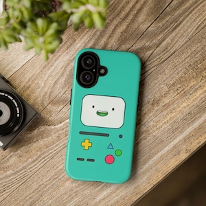 May include: A teal smartphone case featuring a graphic of a rectangular character with a white face, a smiling mouth, a yellow plus sign, a blue triangle, a green circle, and a pink circle, mimicking a handheld video game console design.