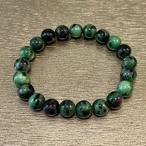 May include: A beaded bracelet composed of round, polished gemstones. The beads are primarily green with dark red and black inclusions. The bracelet is displayed on a light gold surface. The gemstones are approximately 0.4 inches in diameter.
