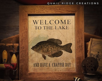 Pode incluir: Uma impressão emoldurada com o texto "WELCOME TO THE LAKE" e "AND HAVE A CRAPPIE DAY". A impressão apresenta uma ilustração detalhada de um peixe sobre um fundo de mapa. A moldura é de madeira rústica.