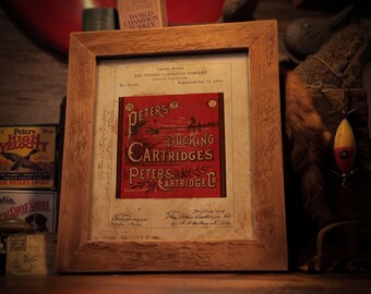 May include: A vintage print of a Peters Cartridge Company advertisement for duck hunting cartridges. The print features a red and white illustration of ducks flying over a lake with the text "25 Peters 19 Ducking Cartridges Peters Cartridge Co. Cincinnati" in black and red lettering. The print is framed in a wooden frame.