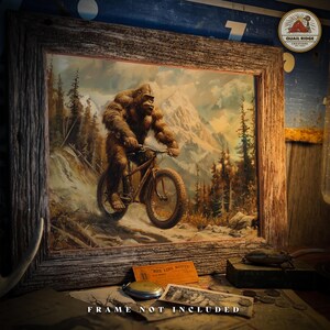 May include: A painting of a Bigfoot riding a mountain bike on a snowy trail in the mountains. The painting is framed in a rustic wooden frame. The text "FRAME NOT INCLUDED" is printed below the painting.