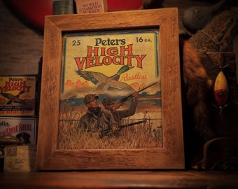 May include: A vintage framed print of a Peters High Velocity 16 GA. shotgun shell advertisement. The print features a hunter aiming a shotgun at a flying duck with the text "Peters High Velocity Rustless" in a yellow banner.