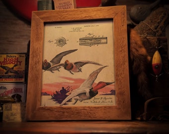 Può includere: Una stampa incorniciata con un'illustrazione vintage di anatre in volo su un lago al tramonto. L'opera d'arte include diagrammi di brevetto e il testo "FRAME NOT INCLUDED". La cornice è in legno marrone chiaro.