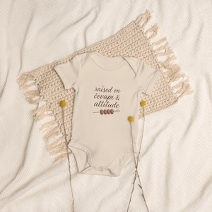 Peut inclure: Body bébé crème avec l'inscription "raised on ćevapi & attitude" en écriture cursive. Un petit graphique de ćevapi sur une brochette est en dessous. Le body est posé sur une couverture beige crochetée.