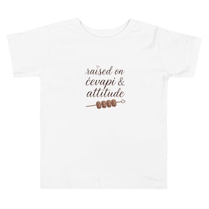 May include: White toddler t-shirt with the text "raised on ćevapi & attitude" in a script font. A graphic of ćevapi on a skewer is below the text. The design is in brown and the heart is pink.