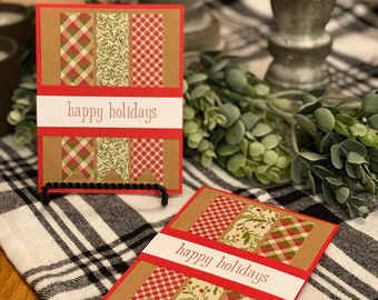 May include: Two handmade holiday cards with a red border and a white banner that says "happy holidays". The cards feature a brown background with red and green patterned paper strips.