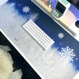 Könnte beinhalten: Eine weiße und blaue Schreibtischunterlage mit einem winterlichen Design mit weißen Bäumen, Schneeflocken und einem sternenklaren Nachthimmel. Die Unterlage liegt auf einem Schreibtisch mit einer weißen Tastatur und einer kleinen Uhr.