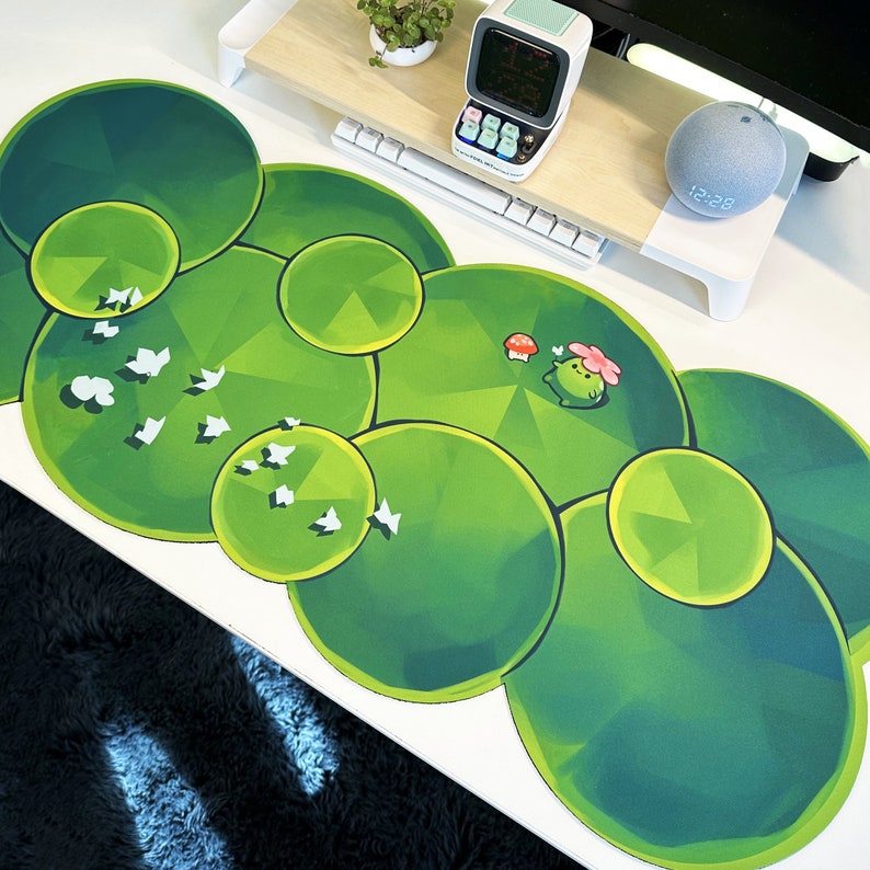 May include: A green and white circular mousepad with a design of overlapping circles and a small frog character. The mousepad is on a white surface with a keyboard and a small clock.