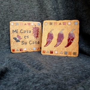 Könnte beinhalten: Zwei Keramikfliesen mit südwestlichem Thema. Eine Fliese zeigt den Text "Mi Casa es Su Casa" mit einem roten Chili-Pfeffer-Design. Die andere Fliese zeigt drei rote Chili-Pfeffer.
