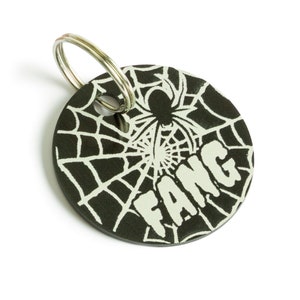 May include: Black metal keychain with a spider web design and the word "FANG" in white lettering.