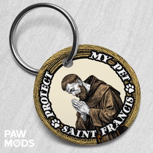May include: A round keychain with a silver ring. The keychain features an illustration of Saint Francis in a brown robe, with the text "PROTECT MY PET SAINT FRANCIS" around the edge. The background is beige and the border is gold.