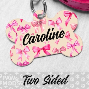 May include: Bone-shaped pet tag with a light beige background and pink bows. The name "Caroline" is written in black script. The tag has a silver ring for attachment. The words "Two Sided" are at the bottom.