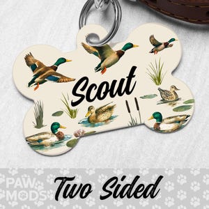 May include: Bone-shaped pet tag featuring illustrations of ducks in a pond setting. The tag is cream-colored with the name "Scout" in black script. The words "Two Sided" are printed at the bottom. A silver ring is attached at the top for easy attachment to a collar.