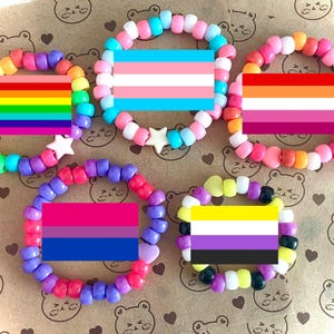 May include: Six colorful beaded bracelets with different pride flags. The bracelets are made of small plastic beads in various colors, including rainbow, pink, blue, purple, yellow, and white. Each bracelet features a different pride flag, including the rainbow pride flag, the transgender pride flag, the lesbian pride flag, the bisexual pride flag, the non-binary pride flag, and the pansexual pride flag.