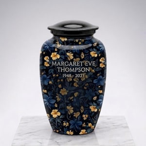 May include: A dark blue cremation urn with a black lid, decorated with gold and blue floral motifs. The urn is inscribed with "MARGARET EVE THOMPSON 1948-2023". It sits on a white marble base.