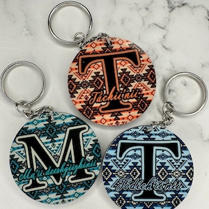 May include: Three round keychains with a black letter and a geometric pattern. The keychains are orange, blue, and white. The letters are "T" and "M". The text on the keychains reads "Tachii'nii" and "olla'ii deeshfhiizhinii".