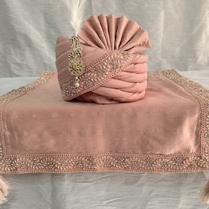 May include: A pink turban with a beaded trim and a gold ornament. The turban is on a pink fabric with a beaded border.