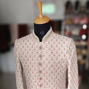 May include: Cream-colored embroidered jacket with a mandarin collar and red accents. The jacket features intricate patterns and decorative buttons. The garment is displayed on a mannequin with a wooden head.