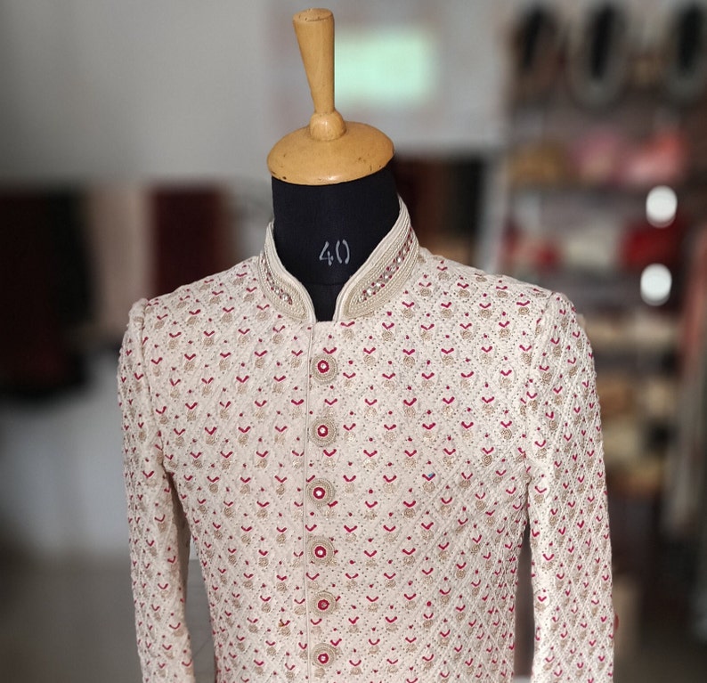 May include: Cream-colored embroidered jacket with a mandarin collar and red accents. The jacket features intricate patterns and decorative buttons. The garment is displayed on a mannequin with a wooden head.