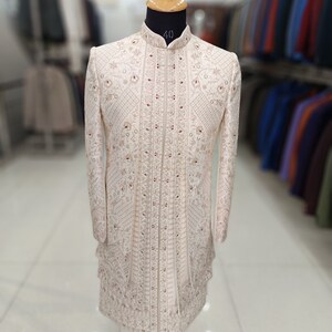 Puede incluir: Una kurta de hombre blanca bordada con cuello alto y adornos rojos. La kurta est&aacute; en un maniqu&iacute; y tiene un dise&ntilde;o tradicional indio.