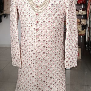 May include: Cream-colored embroidered sherwani with red accents, displayed on a mannequin. The long-sleeved garment features intricate patterns and a mandarin collar. A pearl necklace is draped over the front. The mannequin is on a wooden stand.