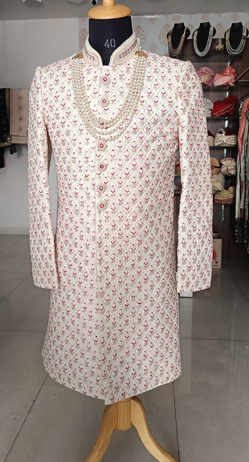 May include: Cream-colored embroidered sherwani with red accents, displayed on a mannequin. The long-sleeved garment features intricate patterns and a mandarin collar. A pearl necklace is draped over the front. The mannequin is on a wooden stand.