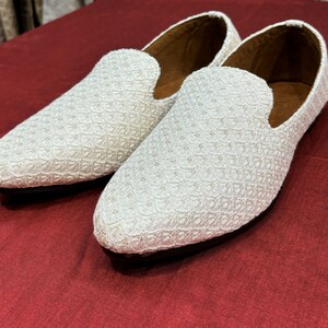 Pode incluir: Um par de mocassins brancos bordados com bico fino. Os mocassins s&atilde;o feitos de um tecido delicado e texturizado e t&ecirc;m um brilho sutil.
