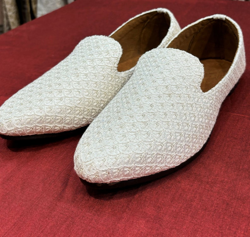 Pode incluir: Um par de mocassins brancos bordados com bico fino. Os mocassins s&atilde;o feitos de um tecido delicado e texturizado e t&ecirc;m um brilho sutil.
