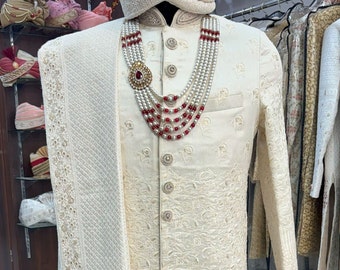 Puede incluir: Un sherwani blanco para hombre bordado con una estola blanca a juego. El sherwani tiene un cuello alto y cierre de botones. La estola está drapeada sobre el hombro izquierdo. El sherwani está decorado con collares de perlas y cuentas rojas.