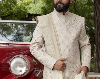 Puede incluir: Un hombre lleva una kurta blanca bordada con un dupatta de color crema drapeado sobre su hombro. Está de pie frente a un coche rojo vintage.