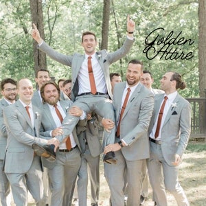Op de afbeelding: Een groep mannen in grijze pakken en oranje stropdassen poseert voor een foto. Eén man wordt door twee anderen omhoog gehouden. De tekst "Golden Attire" is zichtbaar op de achtergrond.