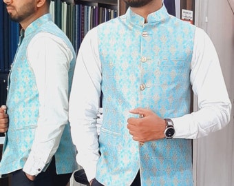 Puede incluir: Un hombre lleva un chaleco azul claro con un estampado floral dorado, una camisa blanca y pantalones negros. El chaleco tiene un cuello mandarín y cierre de botón.