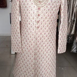 May include: Ivory-colored, long-sleeved sherwani with intricate red and gold embroidery. The sherwani features a mandarin collar and is adorned with a pearl necklace. It is displayed on a mannequin.