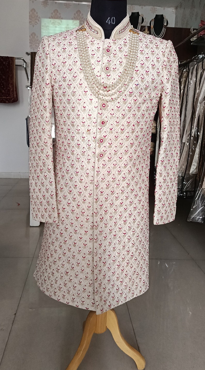May include: Ivory-colored, long-sleeved sherwani with intricate red and gold embroidery. The sherwani features a mandarin collar and is adorned with a pearl necklace. It is displayed on a mannequin.