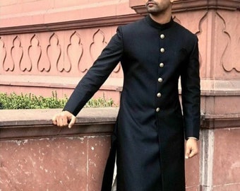 May include: A black long-sleeved kurta with a high collar and gold buttons. The kurta is worn by a person standing in front of a pink wall.