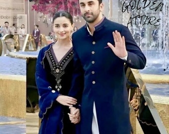 May include: A couple wearing traditional Indian attire. The woman is wearing a dark blue embroidered kurta with a matching dupatta. The man is wearing a dark blue long kurta with a white undershirt and white pants. The text "GOLDEN ATTIRE" is superimposed on the image.