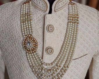 Könnte beinhalten: Eine weiße Perlenkette mit einem goldenen Anhänger mit Kristallen. Die Kette ist mehrlagig und hat ein traditionelles Design. Die Kette wird von einer Schaufensterpuppe mit einem weißen bestickten Hemd getragen.