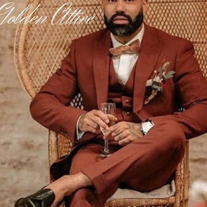 Op de afbeelding: Een man in een driedelig pak in roestkleur met een bruine strik en een corsage. Hij zit op een rieten stoel en houdt een glas champagne vast.