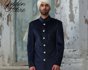 May include: A man wearing a navy blue velvet kurta with gold buttons and a white turban. The kurta has a mandarin collar and long sleeves. The man is standing in front of a white wall with a wooden door behind him.