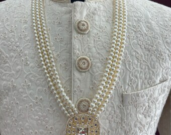 Può includere: Una collana di perle bianche con un ciondolo dorato che presenta un design quadrato con pietre trasparenti. La collana è indossata sopra un capo di abbigliamento bianco ricamato.