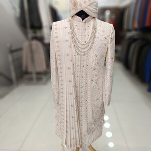 Puede incluir: Un sherwani blanco para hombre bordado con un turbante blanco a juego y un collar de perlas. El sherwani tiene un dise&ntilde;o largo y fluido y est&aacute; decorado con bordados intrincados. El turbante tambi&eacute;n est&aacute; decorado con bordados y tiene una borla dorada. El collar est&aacute; hecho de perlas y se lleva alrededor del cuello. El sherwani se exhibe en un maniqu&iacute; en una tienda.