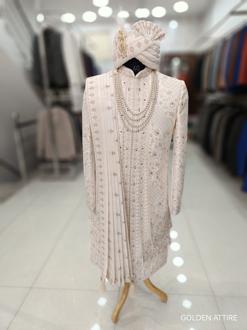 Puede incluir: Un sherwani blanco para hombre bordado con un turbante blanco a juego y un collar de perlas. El sherwani tiene un dise&ntilde;o largo y fluido y est&aacute; decorado con bordados intrincados. El turbante tambi&eacute;n est&aacute; decorado con bordados y tiene una borla dorada. El collar est&aacute; hecho de perlas y se lleva alrededor del cuello. El sherwani se exhibe en un maniqu&iacute; en una tienda.