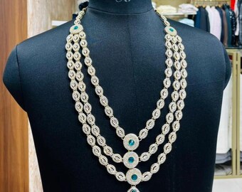 Puede incluir: Un collar de tres hilos con una cadena dorada y piedras redondas transparentes. El collar presenta tres piedras redondas de color verde azulado y un gran colgante de piedra en forma de lágrima de color verde azulado. El collar se exhibe en un maniquí negro.